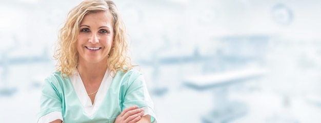 Blonde female doctor smiling into the camera, arms crossed on her chest, dressed in light green uniform with a blurred laboratory background, in a banner format.