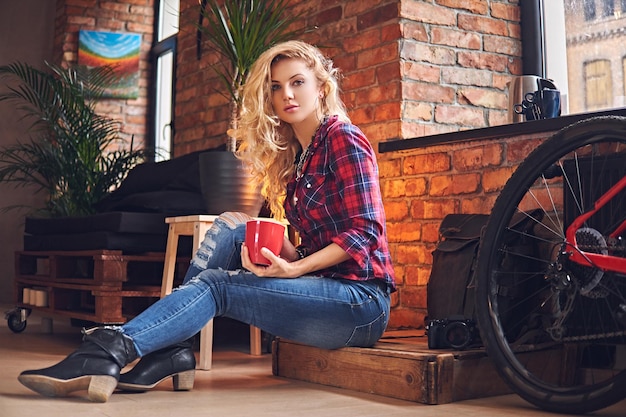 Blonde female dressed in a jeans and fleece shirt, drinks hot coffee near the window in a room with loft interior.