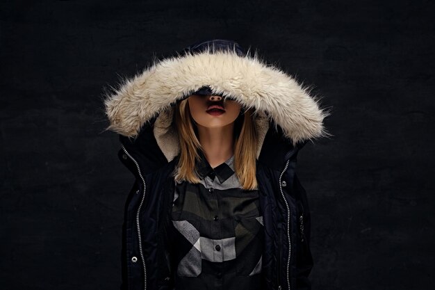 blonde female dressed in winter jacket with deep fur hood isolated on dark background.