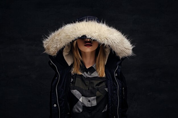 Blonde female dressed in winter jacket with deep fur hood isolated on dark background.