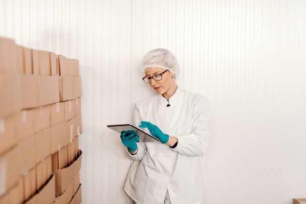 Impiegato femminile biondo in uniforme sterile facendo uso della compressa per logistico mentre stando accanto alle scatole nella fabbrica dell'alimento.