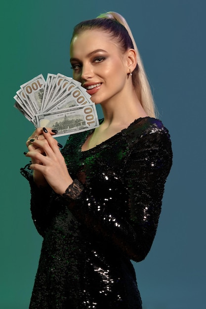 Blonde female in jewelry and black sequin dress smiling showing some hundred dollar bills posing on ...