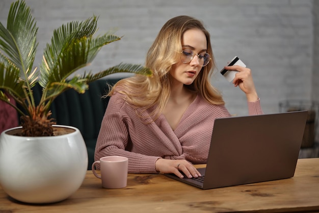 Blonde female in pink cardigan and glasses. Working on her laptop and showing white plastic card. Sitting at wooden table with pink cup, palm in pot on it. Student, blogger. Home interior. Close up