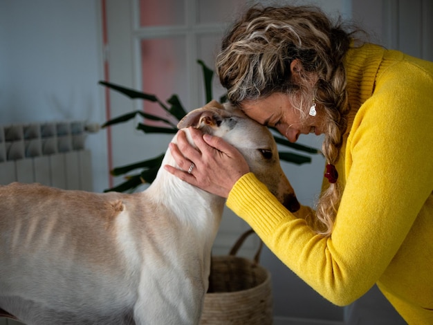 Photo blonde female in a yellow sweater playing with her greyhound dog at home