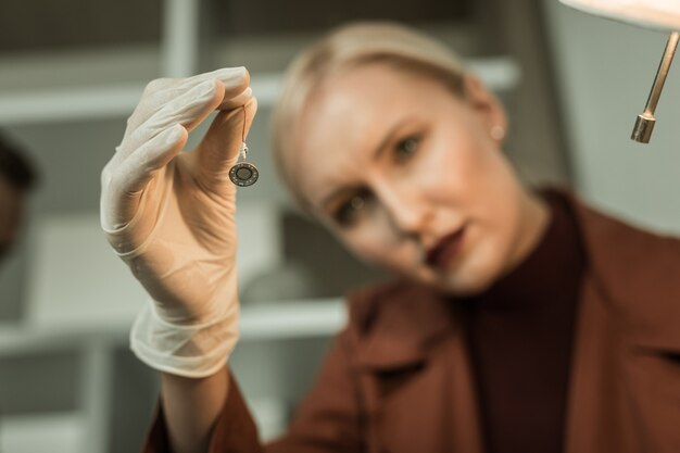 Scienziato forense biondo. donna bionda con rossetto rosso che osserva attentamente il microfono segreto durante la ricerca seria negli appartamenti