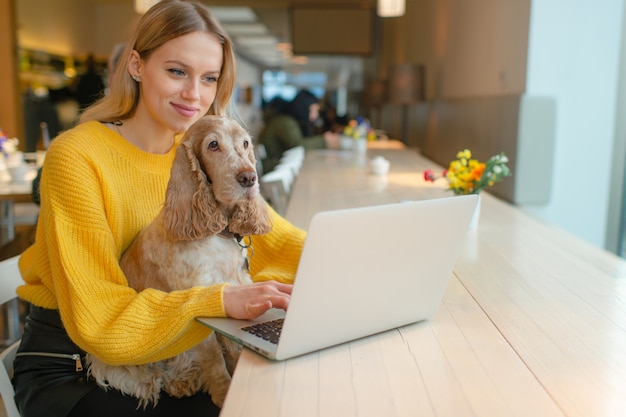 Blonde freelancer blogger in geel sweatshirt met laptop in co-werkruimte en houdt haar oude cocker spaniel hond op haar knieën