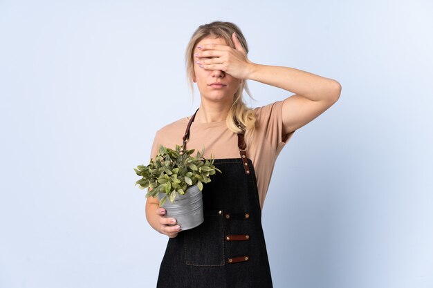 Blonde gardener woman holding a plant over isolated background covering eyes by hands