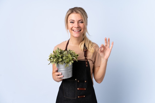 Blonde gardener woman holding a plant over isolated background showing an ok sign with fingers