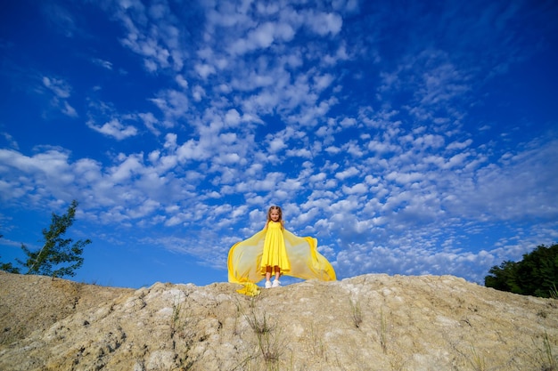 A blonde girl of 7-8 years old in a bright yellow dress against a blue sky.