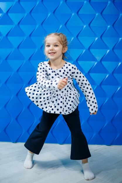 A blonde girl of 7 years old in a jacket with peas on a blue background dances and laughs, bright children's emotions of joy, a happy childhood