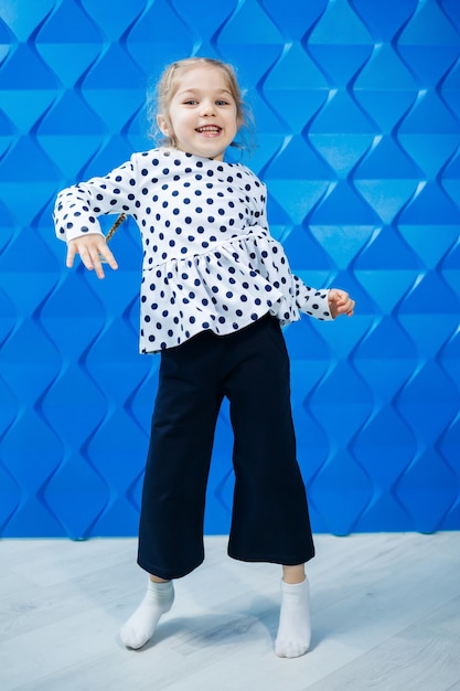 A blonde girl of 7 years old in a jacket with peas on a blue background dances and laughs, bright children's emotions of joy, a happy childhood