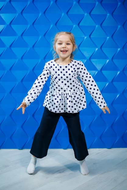 A blonde girl of 7 years old in a jacket with peas on a blue background dances and laughs, bright children's emotions of joy, a happy childhood