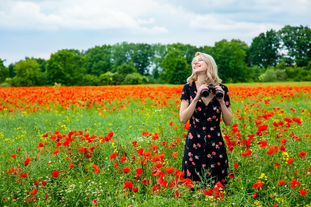 Blonde girl in beautiful dress with binoculars in poppies field in summer time