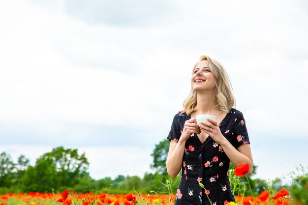 Blonde girl in beautiful dress with cup of coffee in poppies
field in summer time