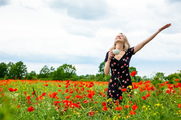 Blonde girl in beautiful dress with cup of coffee in poppies field in summer time