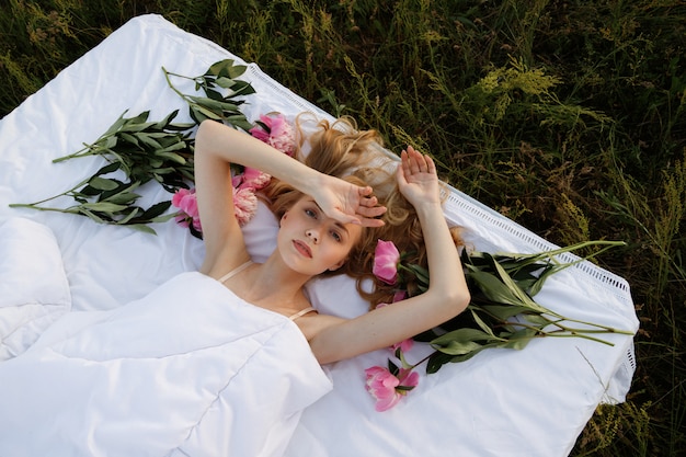 Blonde girl in bed in a field around girl peonies