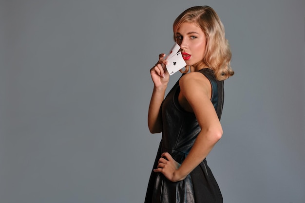 Blonde girl in black dress showing two playing cards posing\
sideways against gray background gambling entertainment poker\
casino closeup