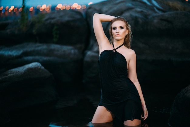Blonde girl in a black dress with blue eyes on the stone coast of the river