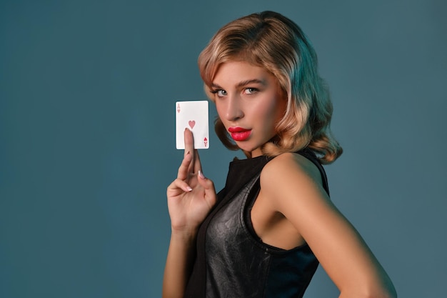Blonde girl in black leather dress showing ace of hearts posing\
against blue background gambling entertainment poker casino\
closeup