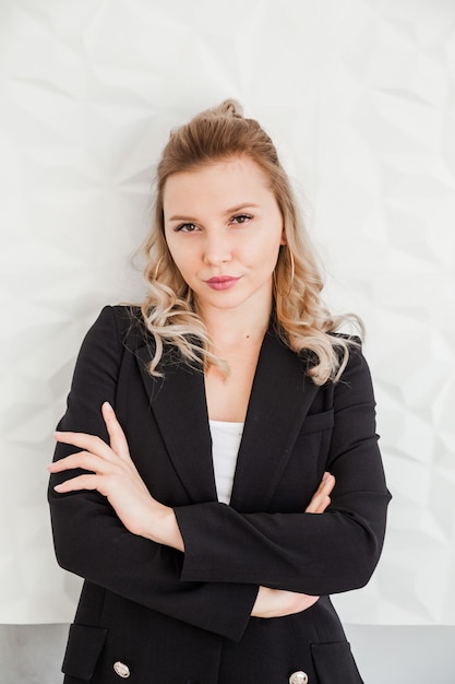 A blonde girl in a black suit stands against a white wall with crossed arms and looks into the camera. Vertycal photo