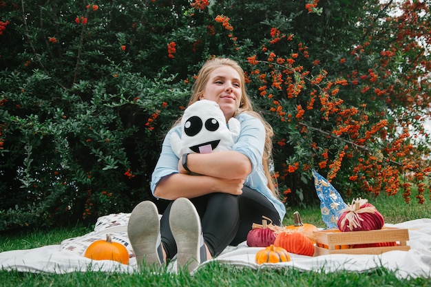 Blonde girl on the blanket in the park on the lawn with a ghost and pumpkins Halloween symbols