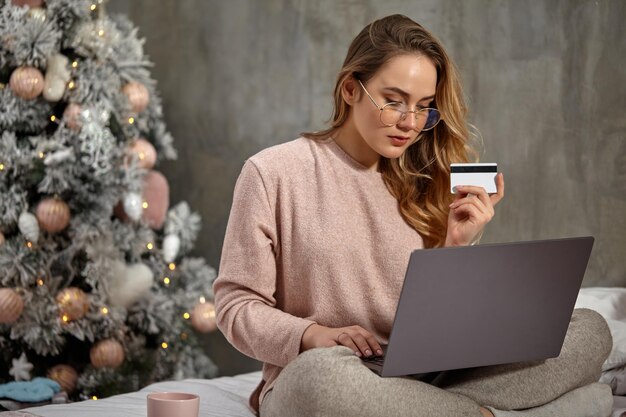 Blogger ragazza bionda con gli occhiali abbigliamento casual tiene in mano una carta di plastica e lavora al suo laptop...