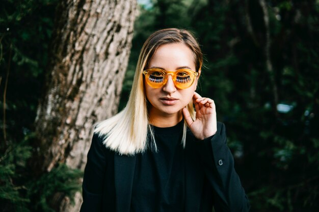 Blonde girl dressed in black jacket with yellow glasses with false eyelashes