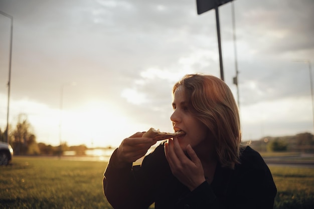 Blonde girl eating pizza sitting outside at sunset