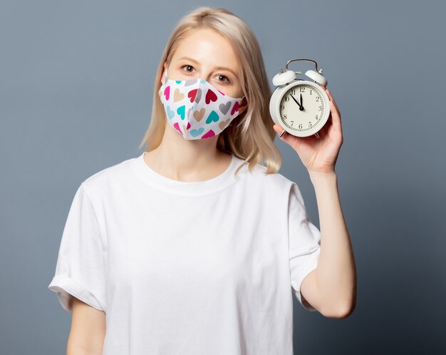 Blonde girl in face mask with with alarm clock on grey space