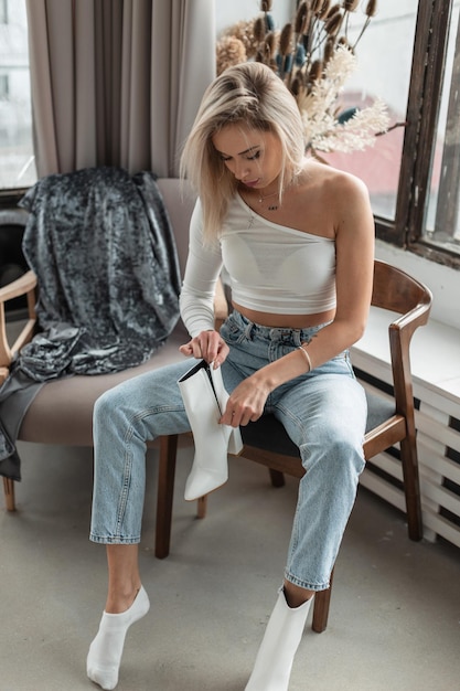 Blonde girl in a fashionable white Tshirt with blue jeans sits on an vintage armchair and puts on leather shoes