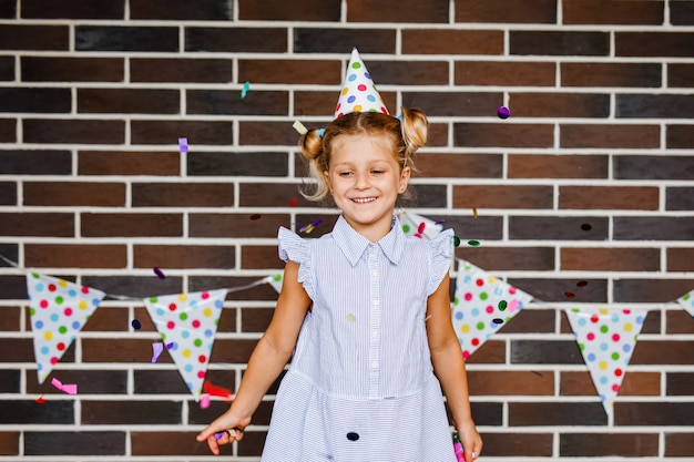 A blonde girl in a festive paper hat laughs and tosses candy on the patio against a brick wall with polka-dot flags.