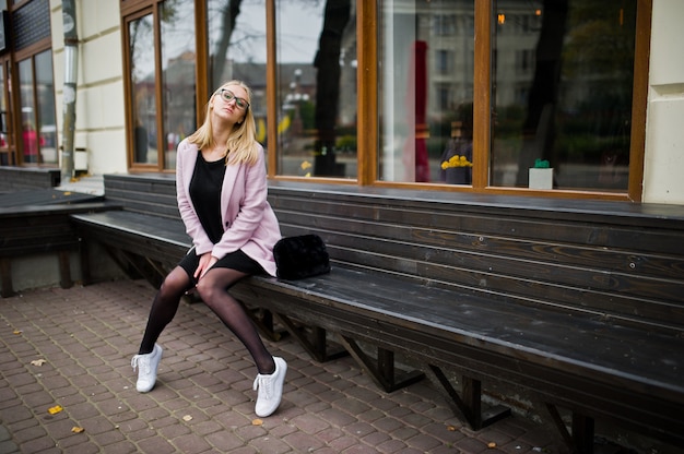 Blonde girl at glasses and pink coat, black tunic sitting on bench at street.