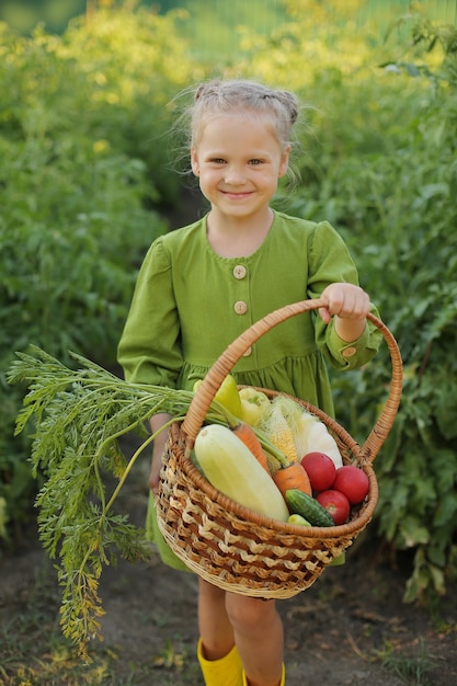 blonde girl in a green dress and yellow boots stands in the garden and holds vegetables picked