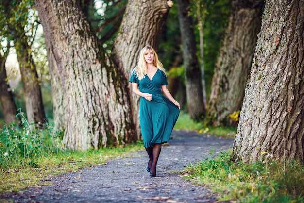 A blonde girl in a green long dress is walking an oak grove