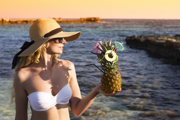 Blonde girl in hat holding a pineapple at the beach at sunset