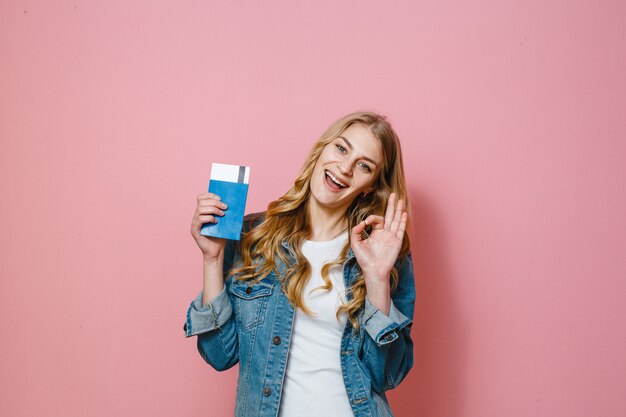 A blonde girl holding a passport with travel tickets and smiling over a pink background