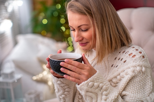 ベッドの上に座ってお茶のカップとセーターでブロンドの女の子