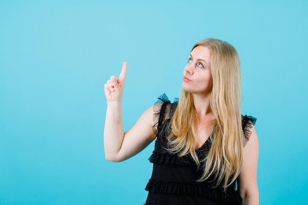 Blonde girl is looking and pointing up with forefinger on blue background