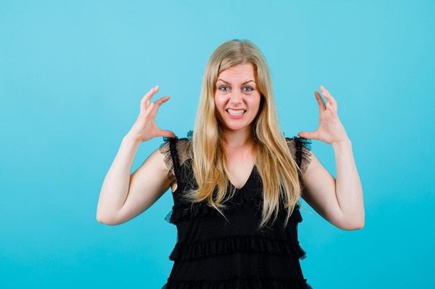 Blonde girl is showing claws gesture by raising up her hands on blue background