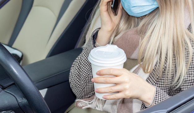 blonde girl is sitting in the car with white eco coffee cup in one hand and phone in another