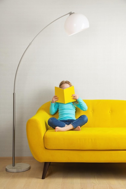 A blonde girl is sitting on the sofa in the room and reading a book in a yellow cover