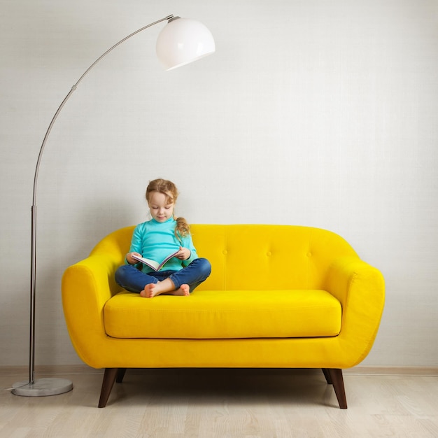 A blonde girl is sitting on the sofa in the room and reading a book in a yellow cover