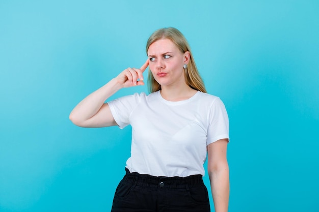 Blonde girl is thinking by holding forefinge ron temple on blue background