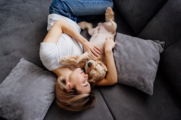 blonde girl in jeans is lying on her back on a gray sofa with a dog