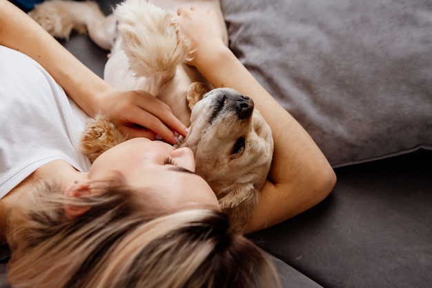 ジーンズのブロンドの女の子は犬と一緒に灰色のソファに仰向けに横たわっています