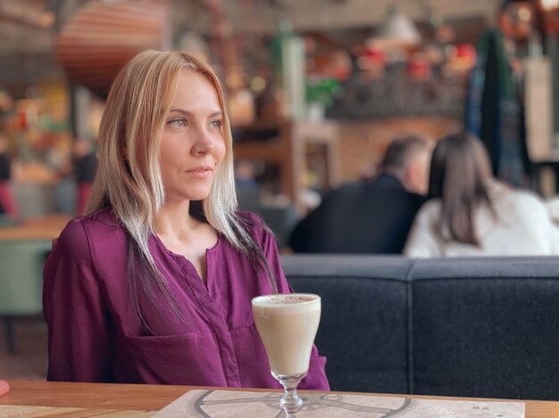 A blonde girl in a lilac sweater sits in a cozy restaurant on a sofa and she is relaxed and smiling and have a coffee drink on her table.