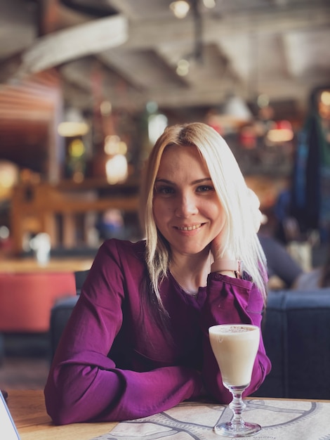 A blonde girl in a lilac sweater sits in a cozy restaurant on a sofa and she is relaxed and smiling and have a coffee drink on her table.