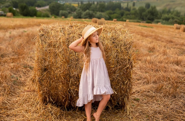 a blonde girl in a pink linen dress and a straw hat stands by a haystack in a mown field