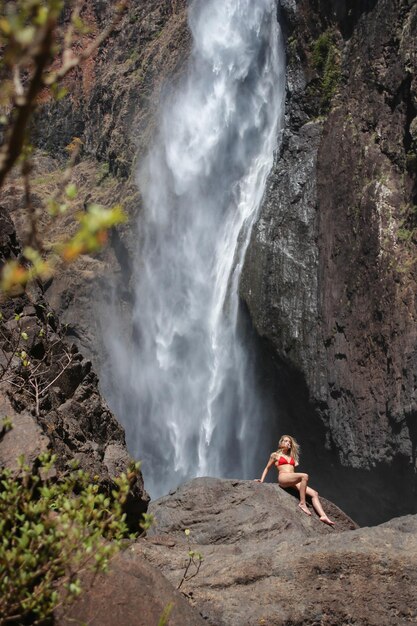 Blonde girl reclining on a large rock under a huge waterfall in Australia