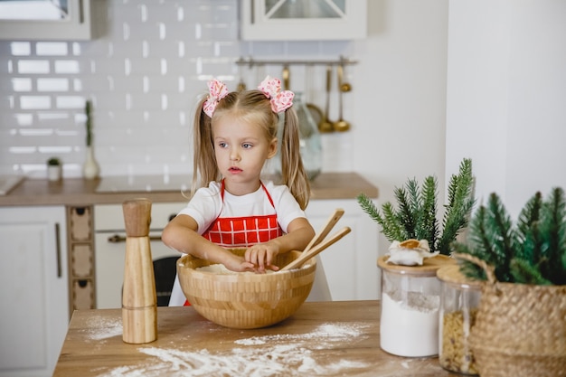 赤いエプロンのブロンドの女の子は、木製のテーブルのキッチンで自宅で生地をこねます